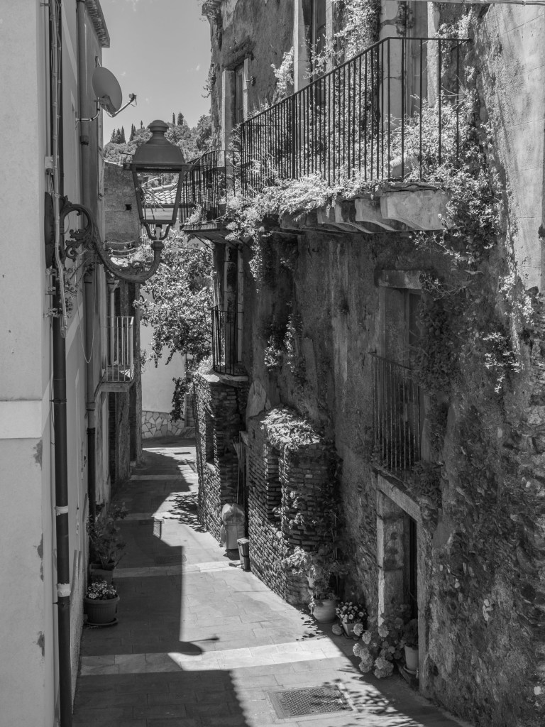 A narrow lane descends between weathered stone walls, balconies, plants and an ornate streetlamp.