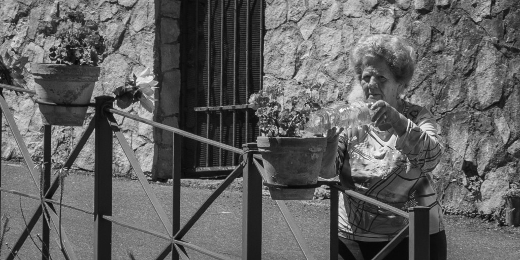 An elderly woman waters potted plants beside a stone wall and metal railing.