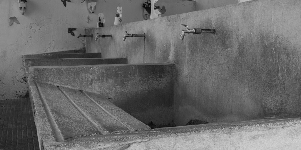 A row of concrete washbasins with metal taps stands beneath small decorative faces and plants.