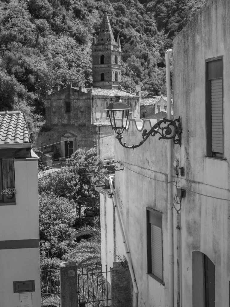 A church tower rises behind houses, trees, cables and an ornate streetlamp in a narrow street.