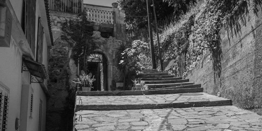 A broad stone staircase climbs between old walls, plants and a weathered arched entrance.
