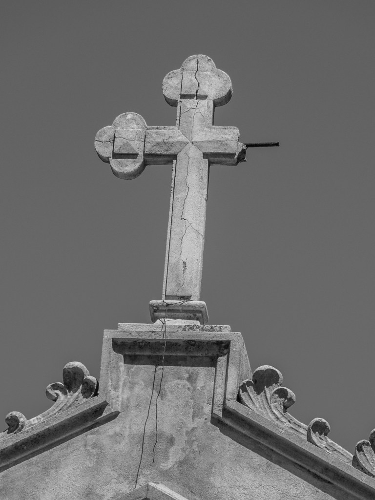 A cracked stone cross rises above the decorated peak of a church façade against a clear sky.