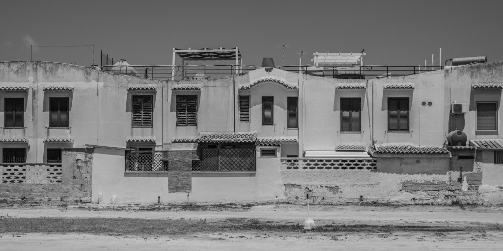 A long row of adjoining houses stands behind a weathered wall and an empty strip of ground.
