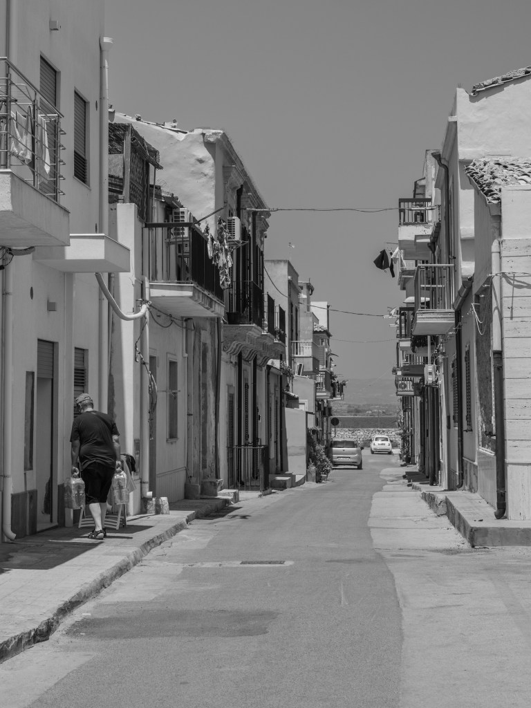 A man carrying large bottles walks along a narrow residential street lined with balconies and low houses.