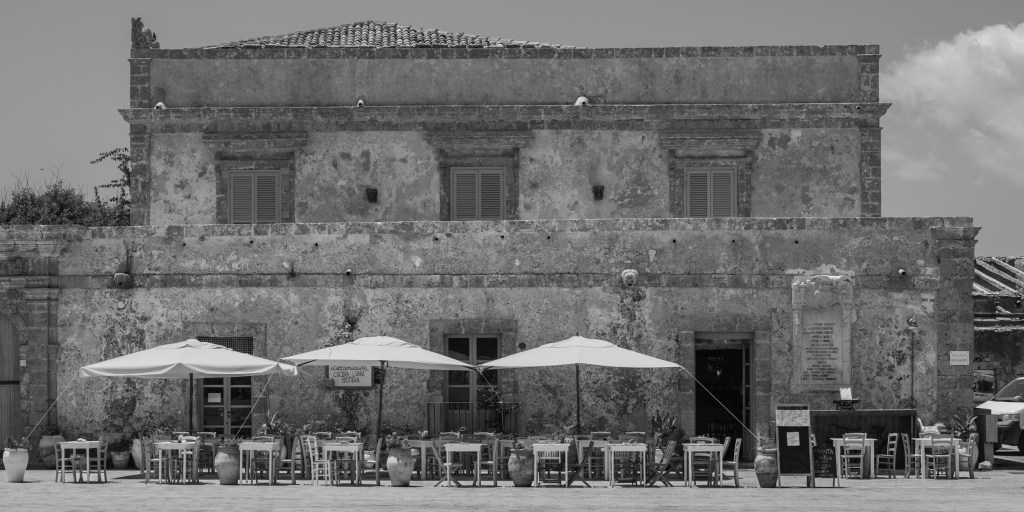 Empty restaurant tables and umbrellas stand before the worn façade of a historic building.