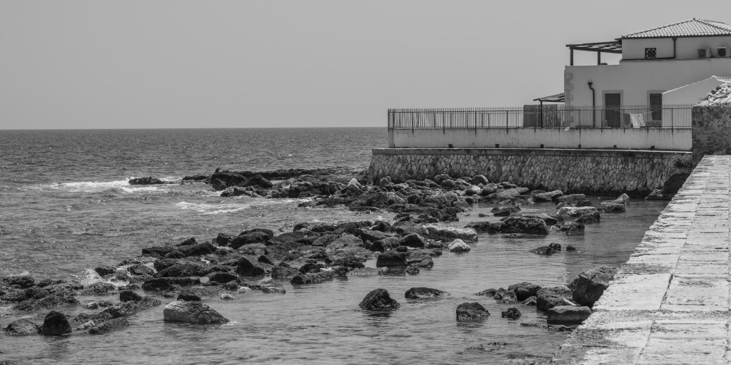 A white waterfront building rises above rocks and shallow water beside a stone walkway.