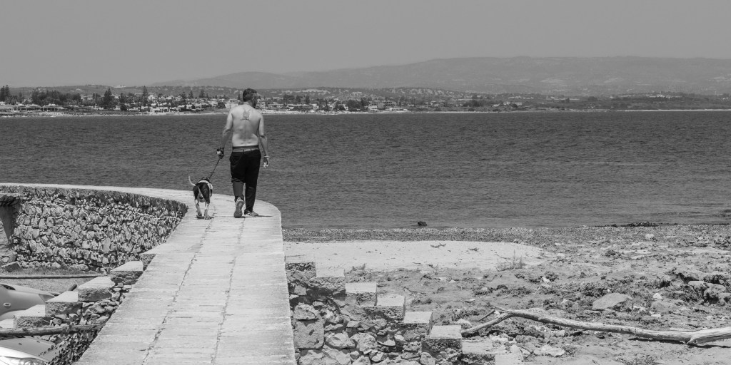 A shirtless man walks a dog along a narrow wall beside the sea, with the distant coast beyond.
