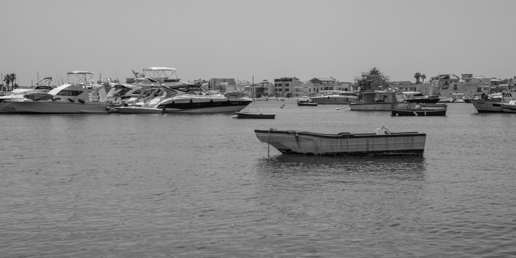 A small open boat floats in the foreground of a harbour filled with fishing boats and luxury motorboats.