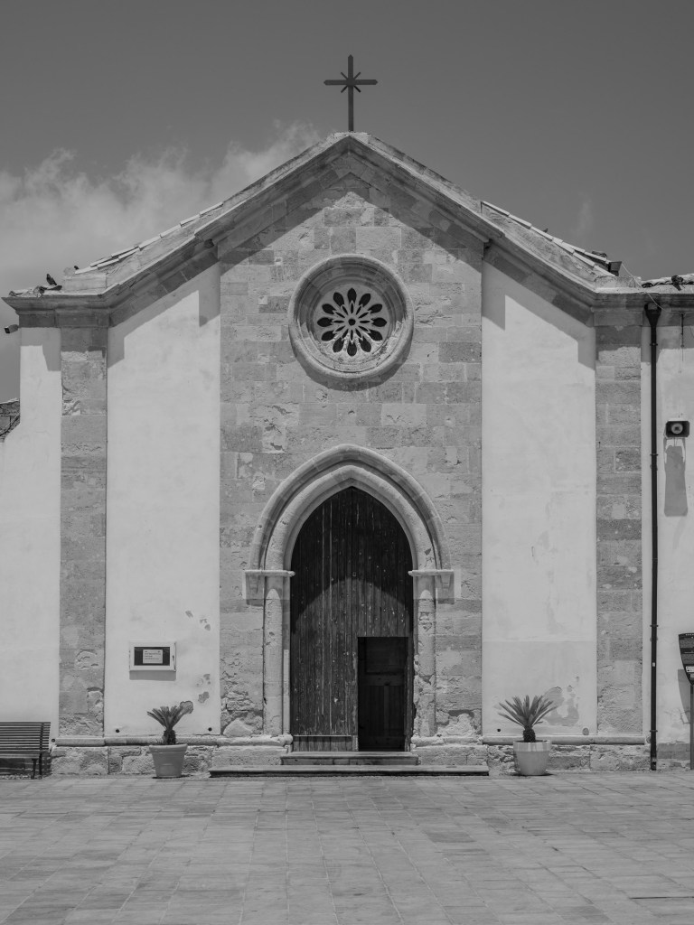 A small church façade has a pointed doorway, circular rose window and cross beneath a clouded sky.
