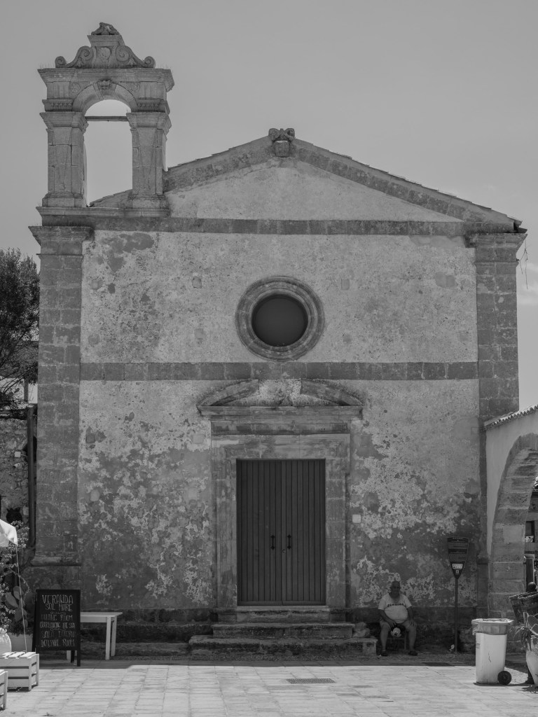 A seated man rests in the shade at the base of a worn church façade with an empty bell opening.