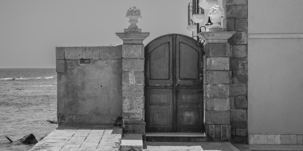 A tall wooden gate between stone pillars stands directly beside the open sea.