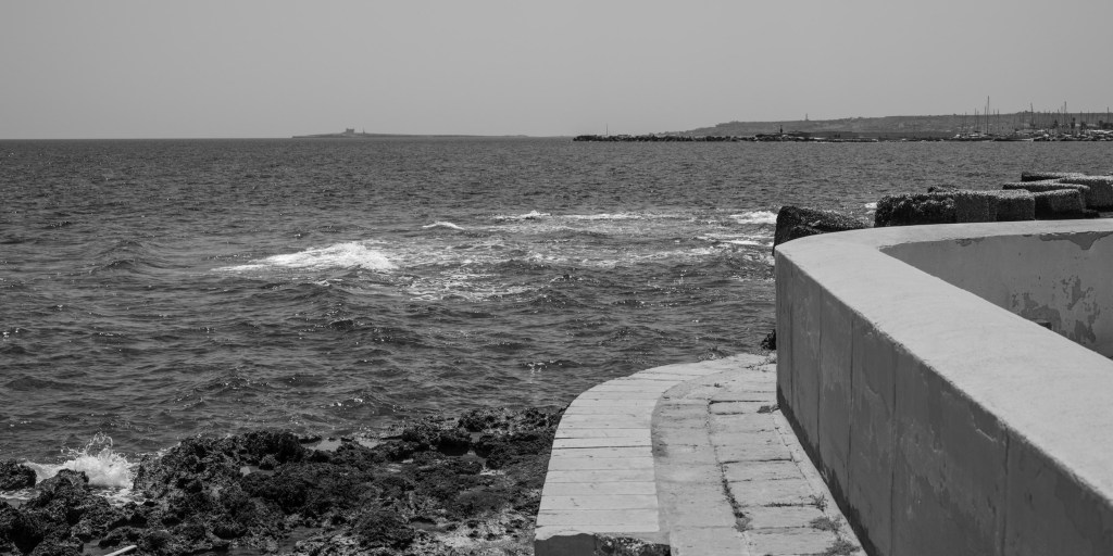 A curved concrete wall and narrow coastal path follow the rocky edge of the sea.