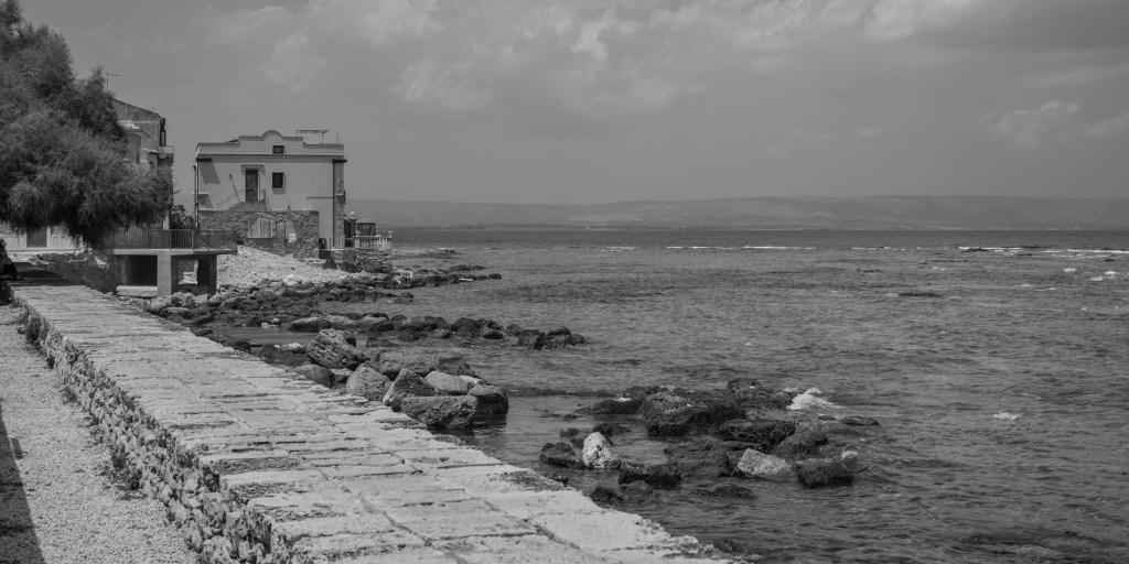 A stone walkway follows a rocky shoreline towards a solitary waterfront house beneath a clouded sky.