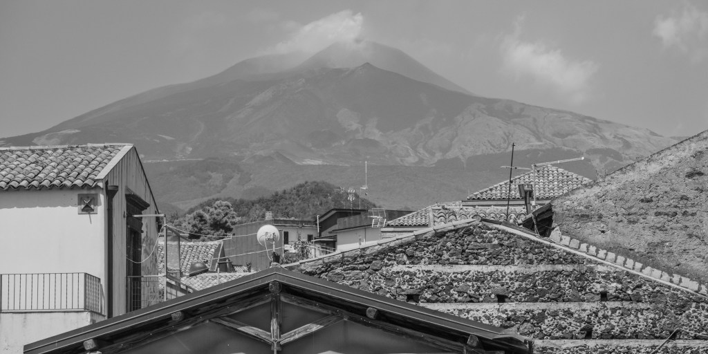 Mount Etna rises behind the tiled roofs and dark volcanic-stone buildings of Nicolosi.