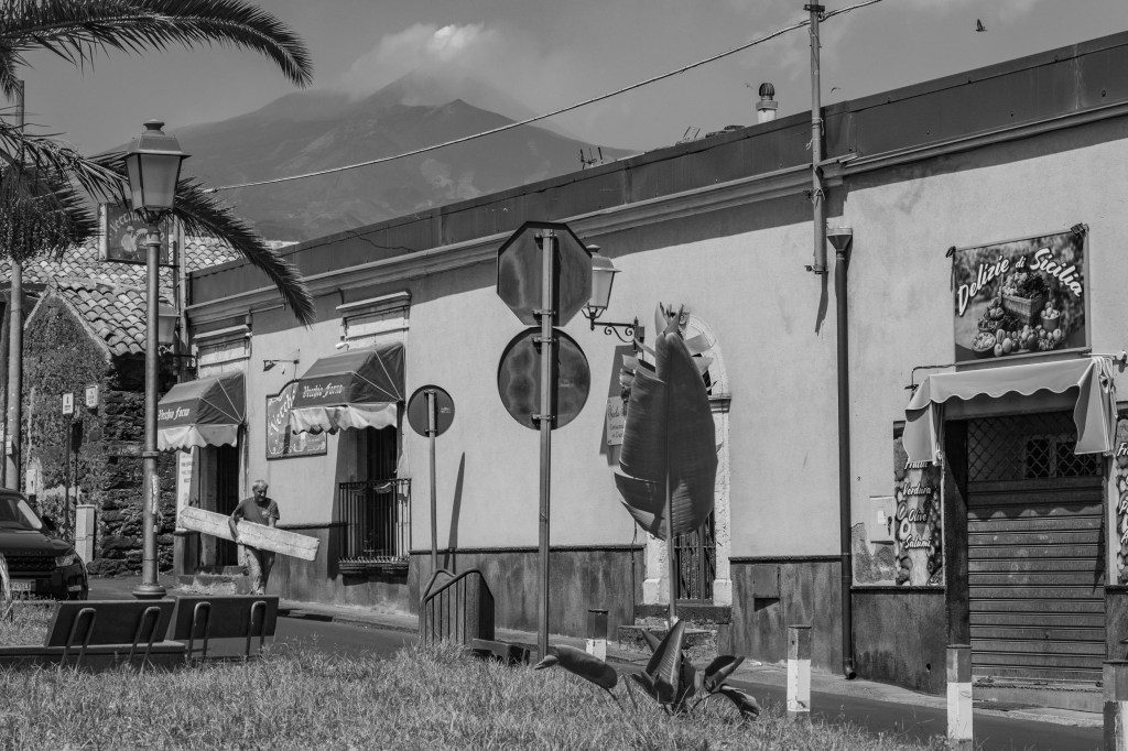 A man carries a long wooden plank past shops and street signs, with Mount Etna rising behind the town.