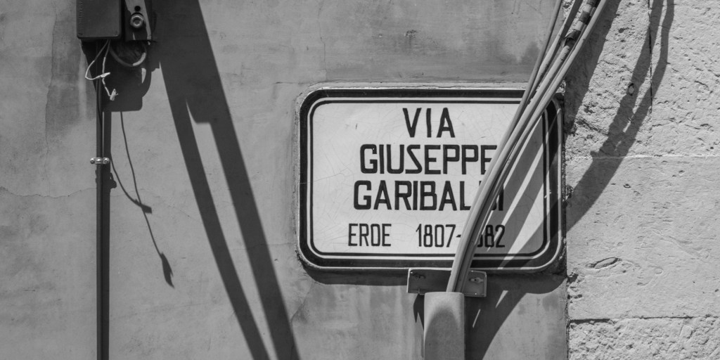 A street sign for Via Giuseppe Garibaldi is partly obscured by pipes and sharp shadows.