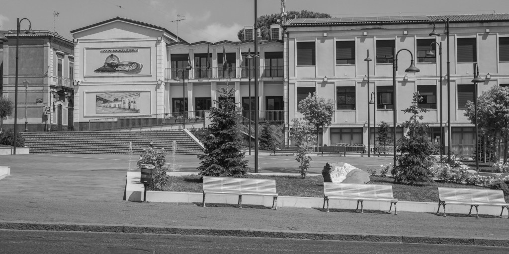 An almost empty civic square contains benches, trees, a small fountain and a large municipal building.