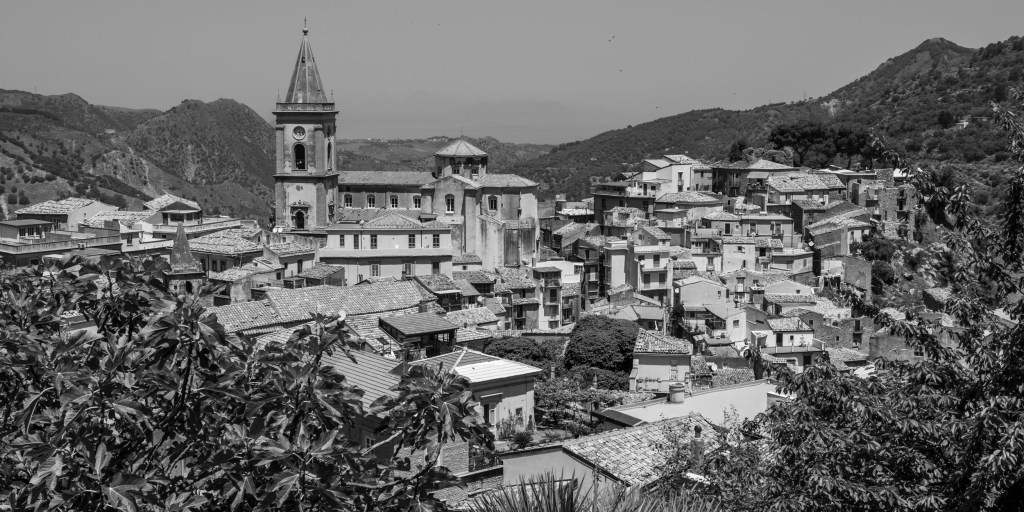 Novara di Sicilia fills a mountain valley, with tiled roofs and a tall church tower surrounded by wooded slopes.
