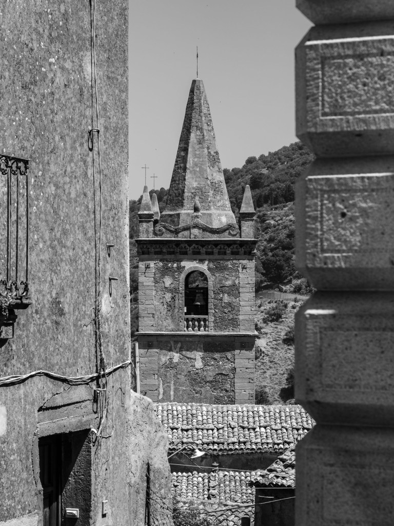 A pointed stone bell tower is framed between weathered buildings, rooftops and a nearby hillside.