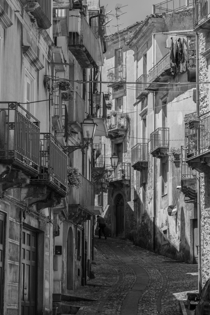 A steep cobbled street climbs between closely packed houses, balconies, laundry and a solitary pedestrian.