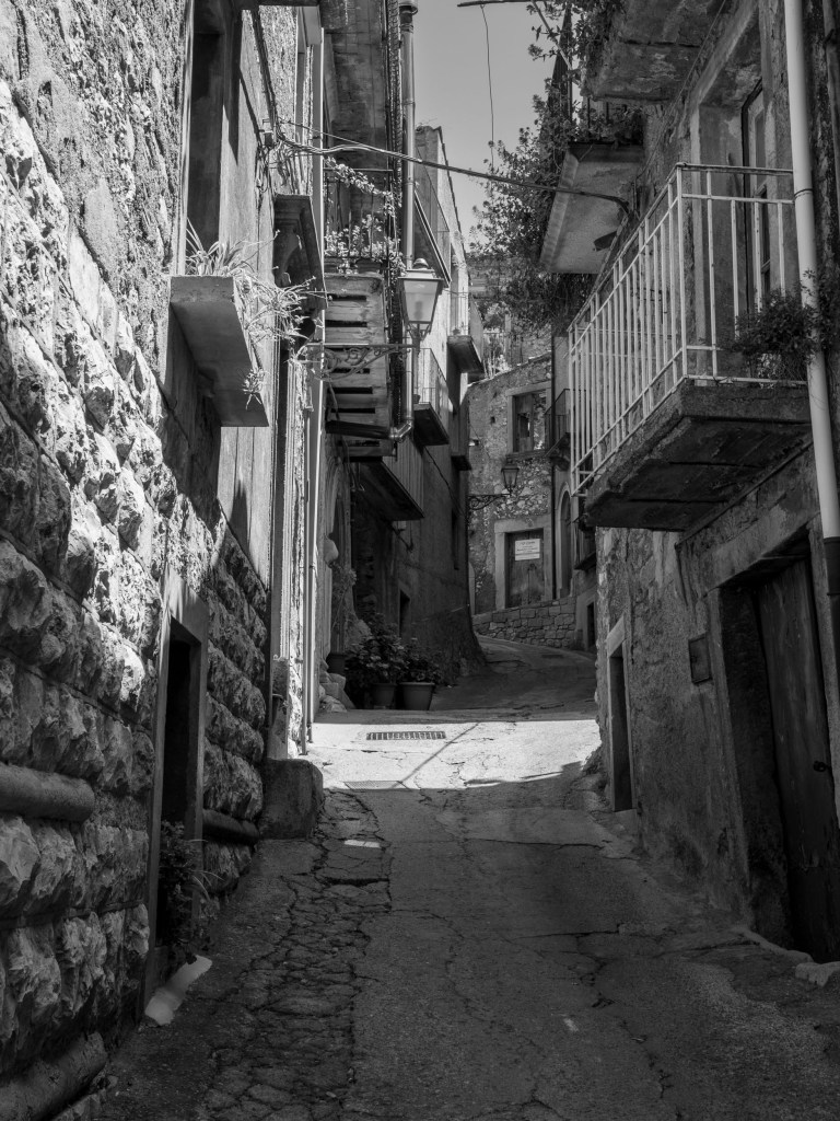 A steep, cracked lane rises between rough stone walls, balconies, plants and overhead cables.