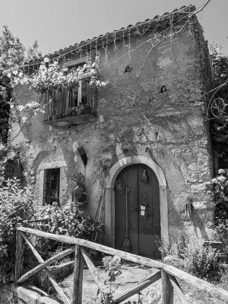 An old stone house with an arched wooden door is surrounded by vines, plants and a rustic timber railing.