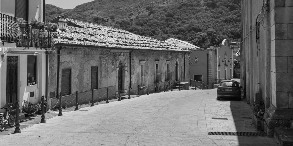 A broad sloping street passes a long weathered building, parked car and mountain hillside.