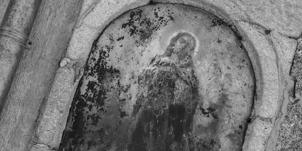 A heavily weathered devotional image of a saint survives within a cracked arched niche.