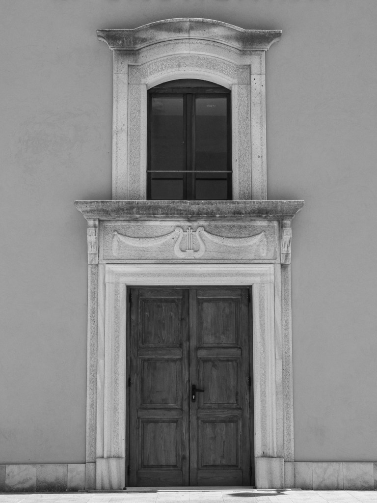 A symmetrical façade has a wooden double door, a dark upper window and a carved lyre motif between them.