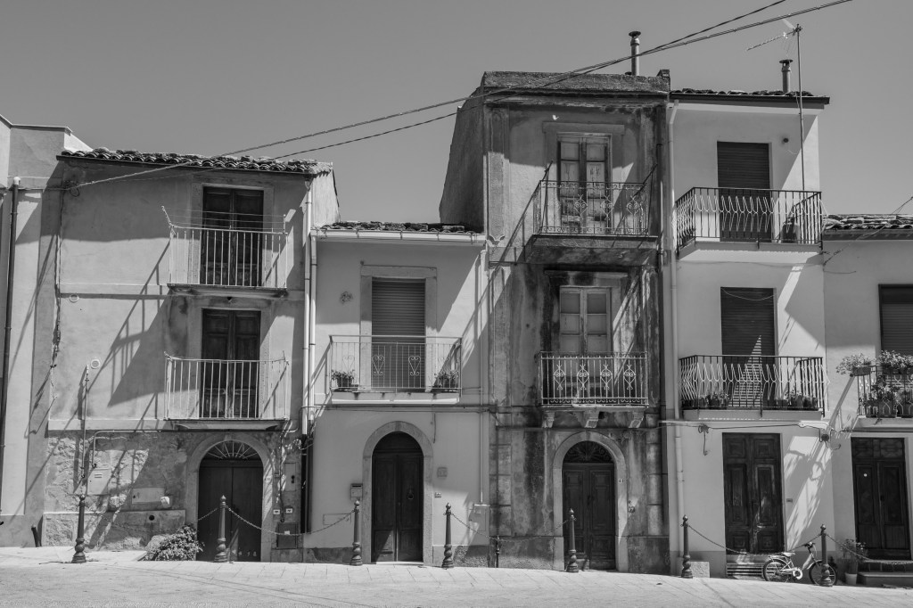 A row of narrow houses with balconies and shuttered doors stands in strong sunlight, with a small bicycle at the far right.