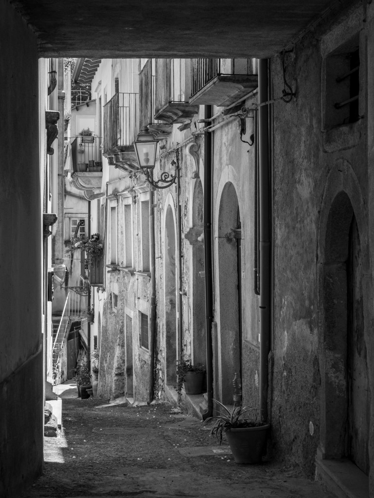 A shadowed covered passage opens onto a narrow street lined with worn façades, balconies and an ornate lamp.