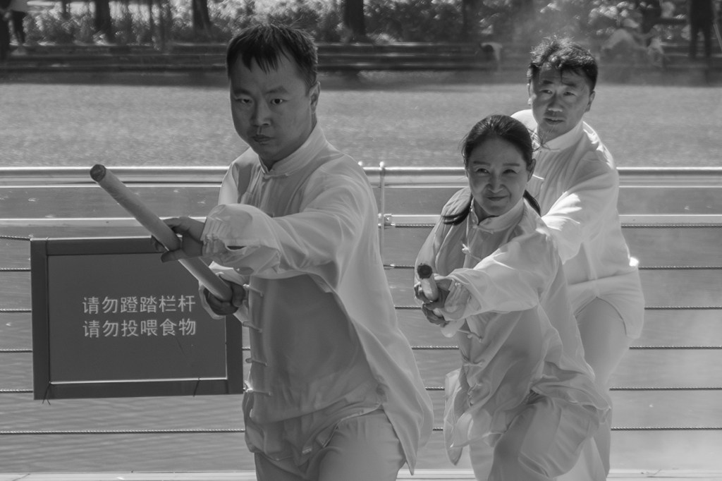 Black-and-white photo of three people in white martial arts uniforms practicing with short wooden sticks on a sunlit wooden platform in a park. They stand in a staggered line, extending their weapons forward, with blurred trees, spectators, railings, and water mist in the background.