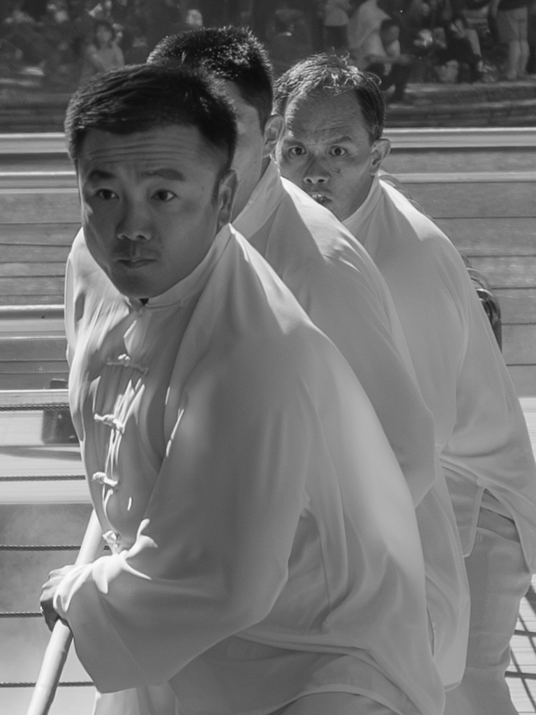 Black-and-white photo of three people in white martial arts uniforms performing a coordinated movement with short wooden sticks. They are aligned diagonally on a wooden deck, partly framed by railings and strong shadows, with park visitors and trees blurred in the background.
