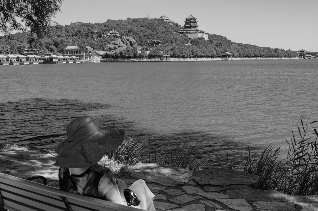 Woman with a large hat sitting on a bench in front of a lake in Beijing.