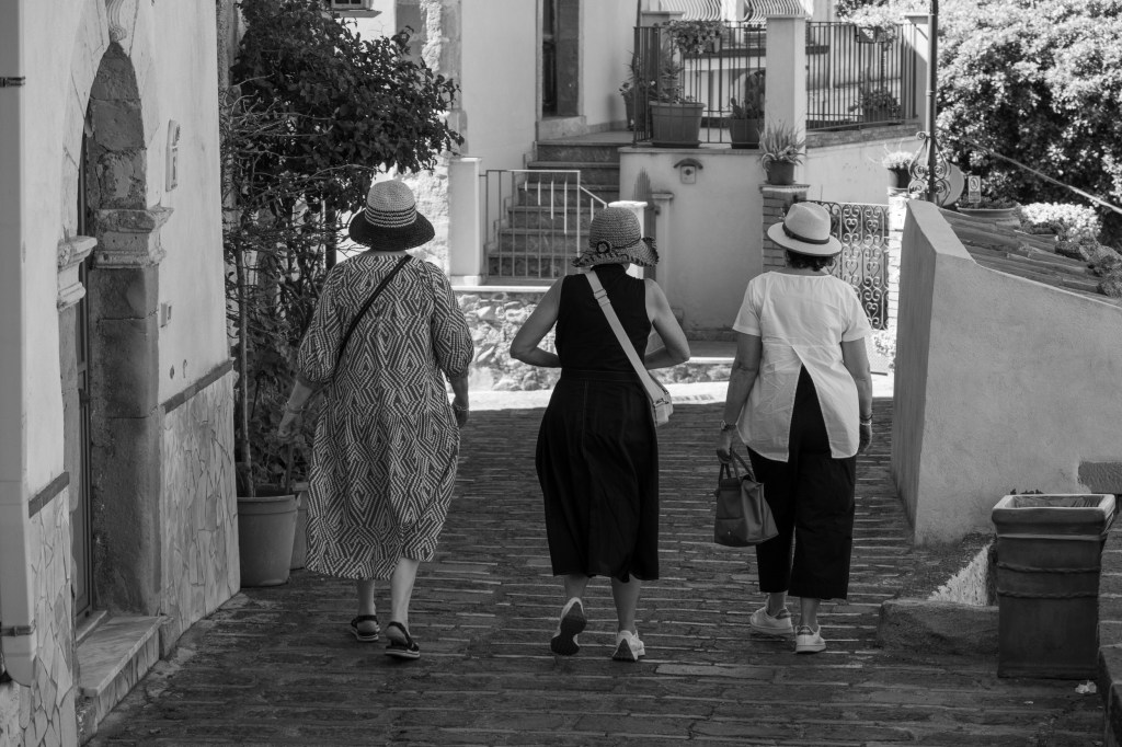 Three women wearing hats walk away along a paved street between white houses and garden walls.