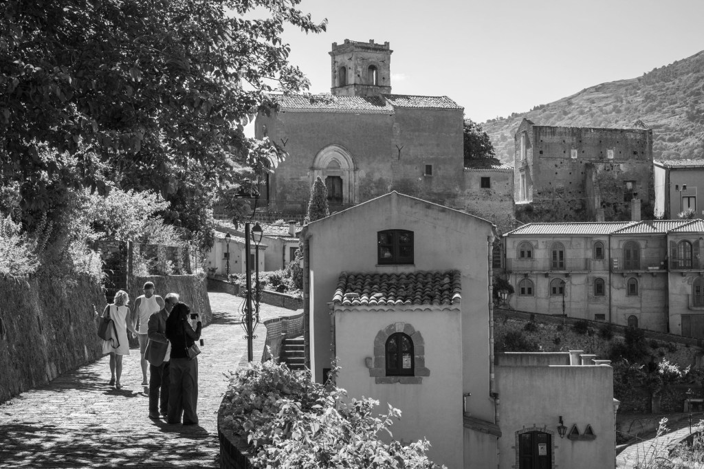 Visitors stand and walk along a sloping street overlooking old churches, houses, and hillside buildings.