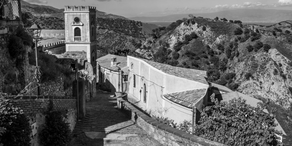 Church and bell tower above a descending road, with steep green hills and a distant coastline beyond.