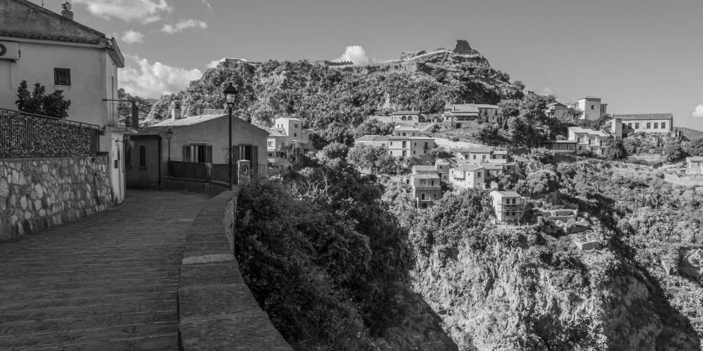 Curving paved road overlooking houses scattered across a rocky hillside beneath the remains of a hilltop structure.