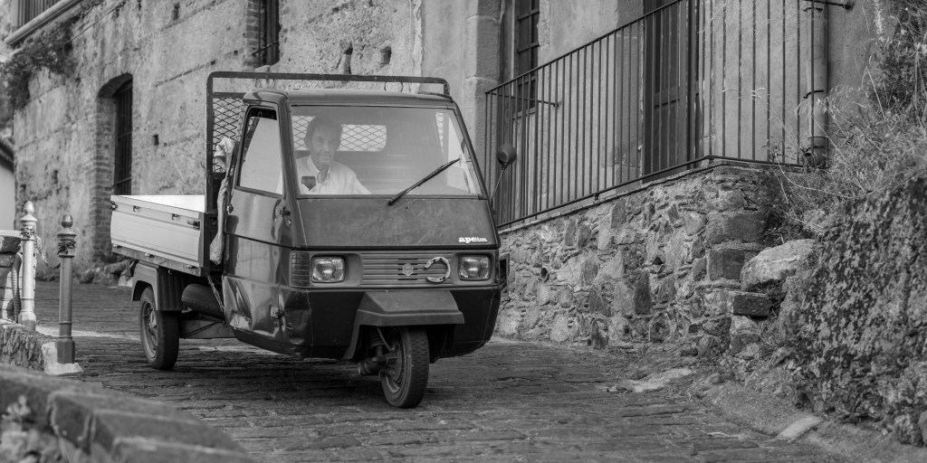 Man driving a small three-wheeled utility vehicle along a narrow stone street beside an old building.