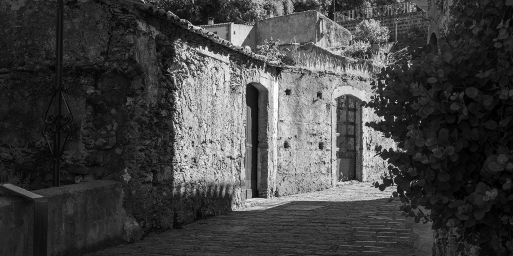 Sunlit ruined wall with arched openings at the end of a shadowed stone lane, partly framed by foliage.