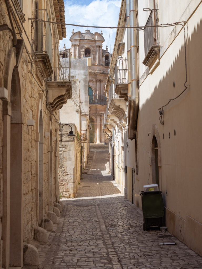 Narrow cobbled street framed by stone façades, balconies, cables, and a Baroque church rising at the far end.
