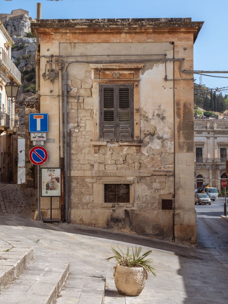 Weathered stone building at a street junction, surrounded by traffic signs, exposed cables, a planter, and passing vehicles.
