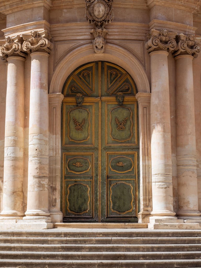 Large faded green wooden church door set between pale stone columns and elaborate carved ornament.