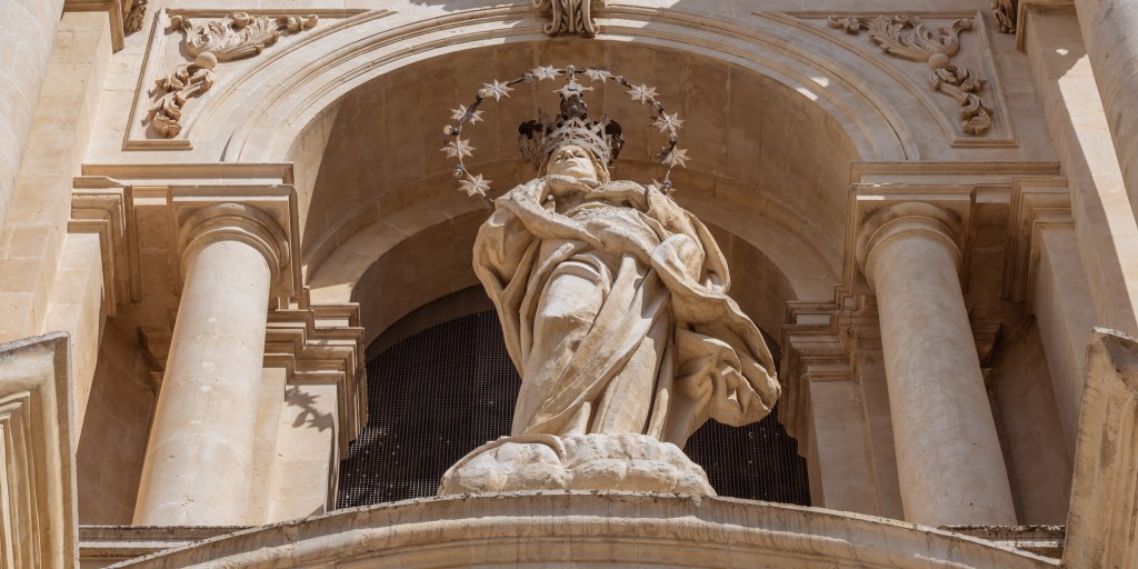 Low-angle view of a crowned stone religious statue beneath an arch, surrounded by columns and a halo of metal stars.