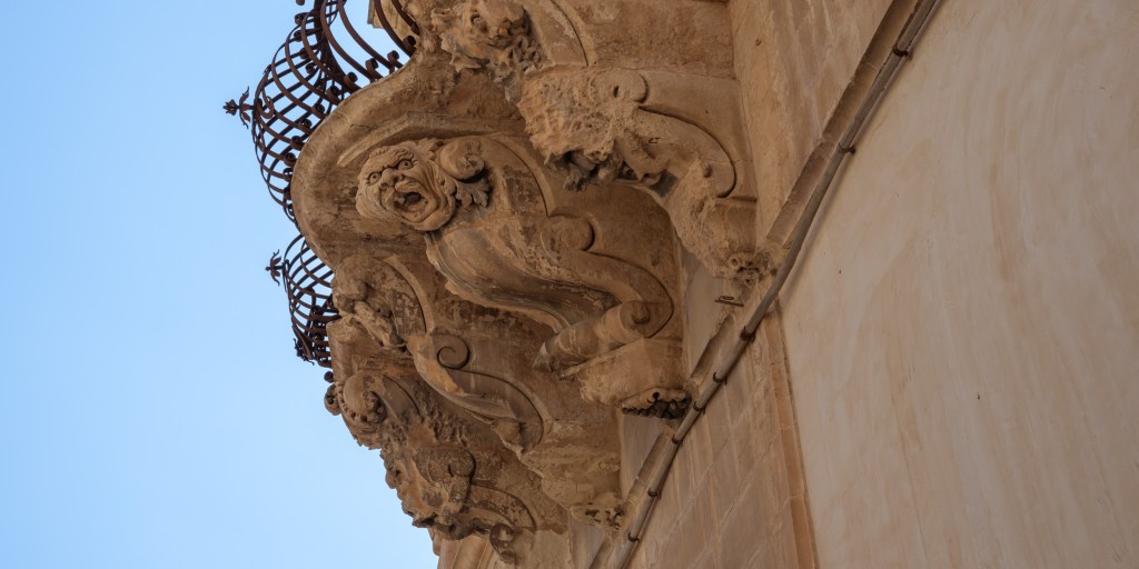 Tilted view of an ornate Baroque façade with a grotesque carved face, curling stonework, iron balconies, and blue sky.