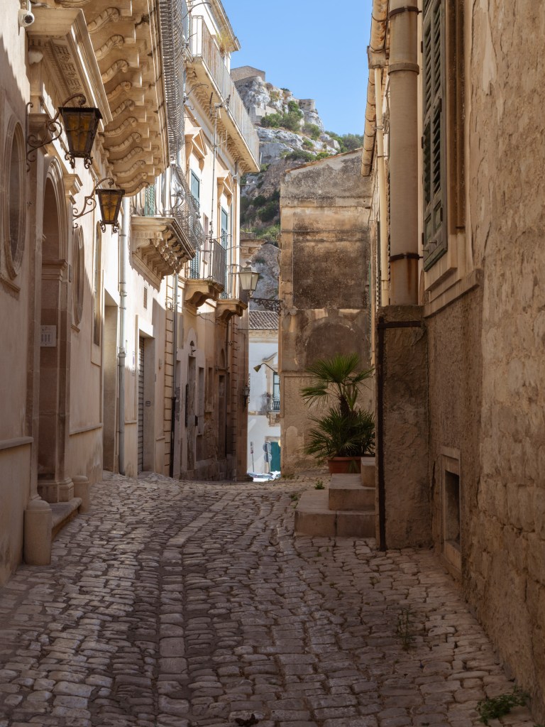 Narrow cobbled street lined with balconies and stone walls, with a rocky hillside visible above the buildings.