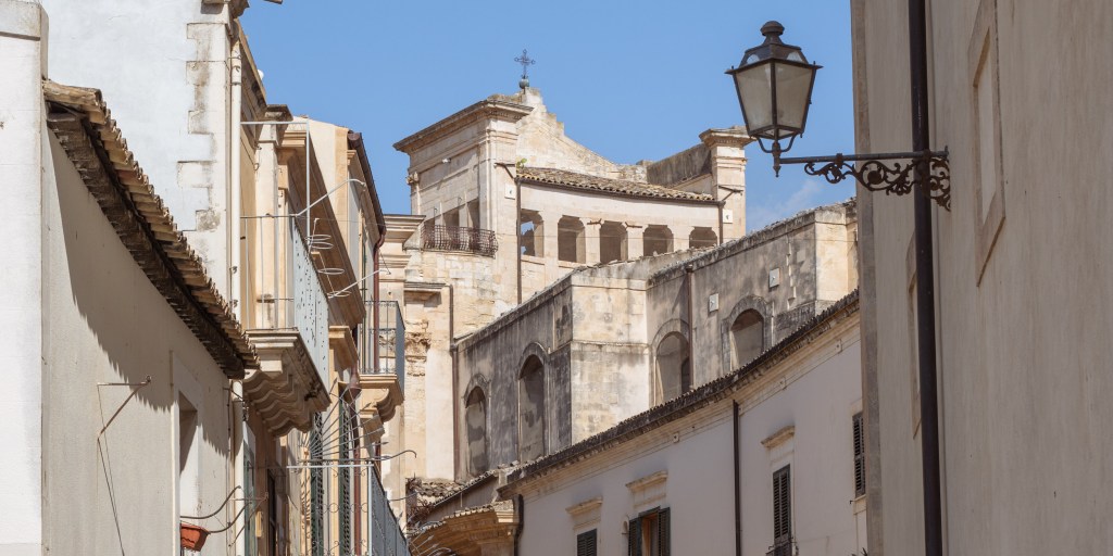 Horizontal street view toward pale stone church buildings, framed by balconies, rooftops, wires, and a hanging streetlamp.