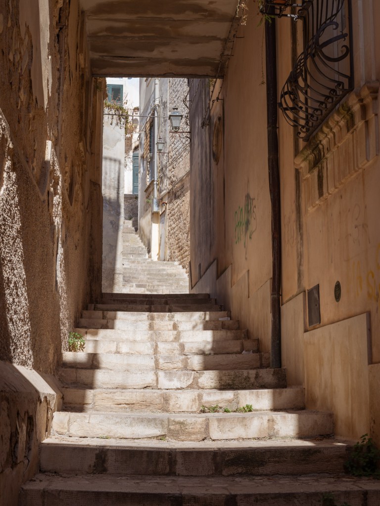 Stone staircase ascending through a shadowed passage, with patches of sunlight, textured walls, ironwork, and distant shutters.