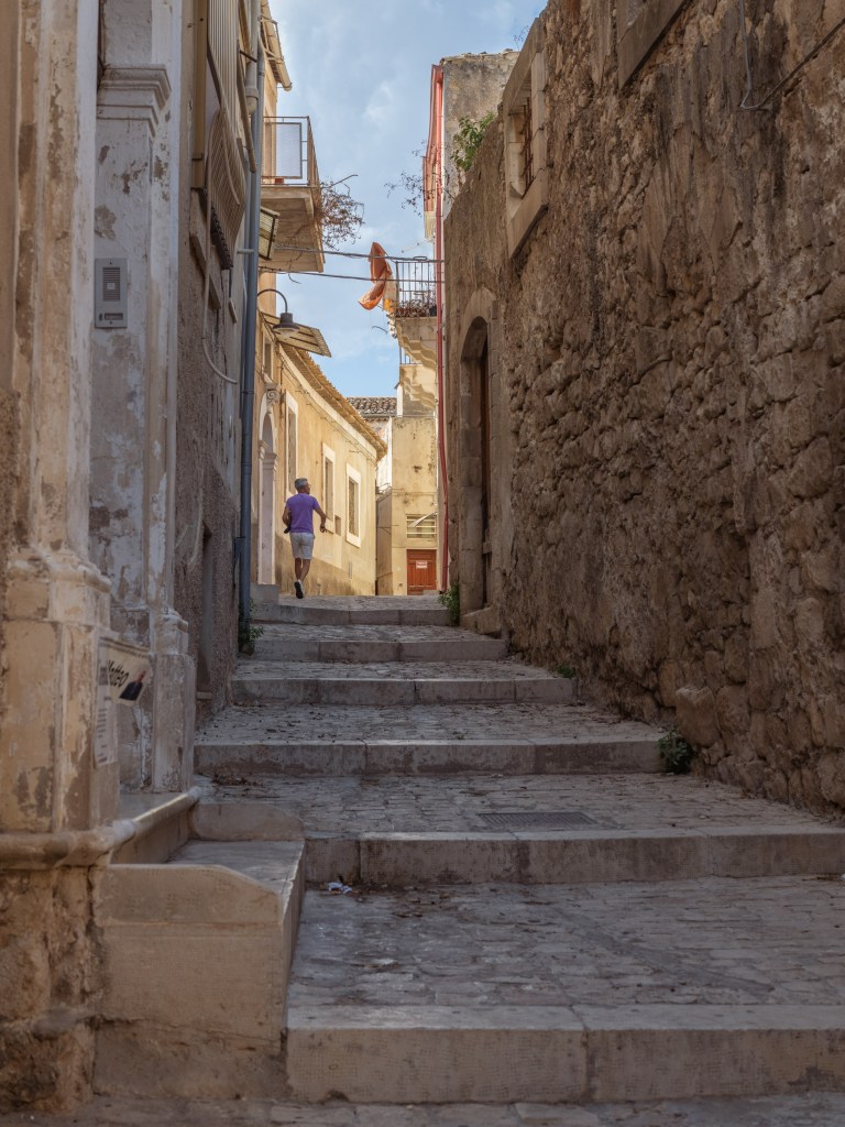 Man in a purple shirt walking away along a stepped stone alley, with an orange cloth hanging from a balcony above.