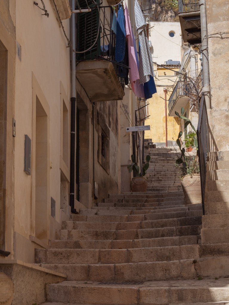 Steep residential staircase lined with balconies, hanging laundry, prickly pear plants, large pots, and weathered walls.